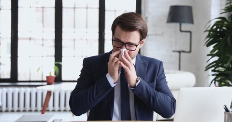 Unhealthy young manager in suit working on computer, feeling first flu symptoms during workday. Unhappy 30s employee in formal suit suffering from runny nose, using paper handkerchief at workplace.