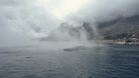 Drone tracking shot of foggy ocean waters and a view of the shoreline, mountains and the beautiful clear skies from afar.