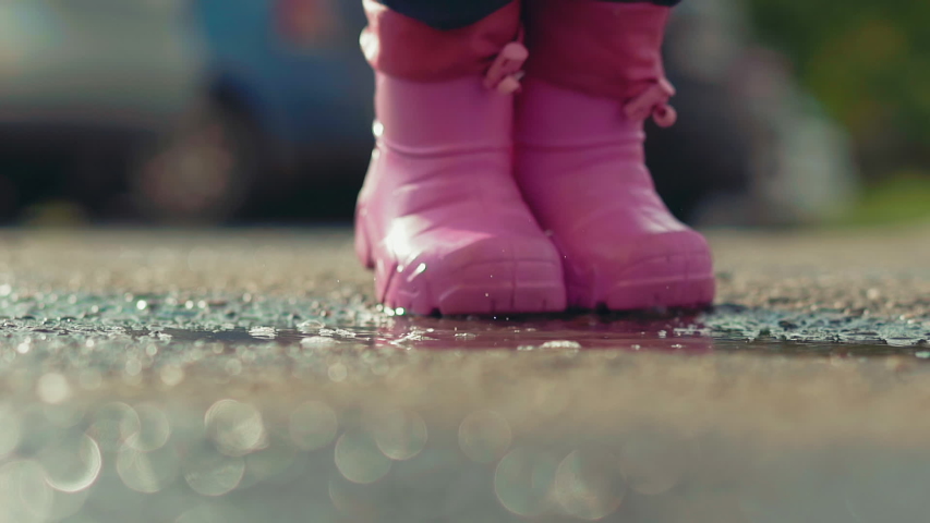 slow motion extreme closeup child in pink rubber waterproof boots jumps into puddle after raining in sunny weather, splashing fun up and out of side of fountain in green park in summer