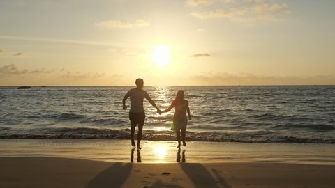 silhouettes of joyful couple running along beach joining hands against ocean waves at tropical sunset slow motion