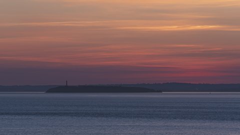 Sunset timelapse. Vibrant orange sky, misty clouds flowing. Island with Lighthouse in far horizon. Large sun telephoto zoom. Cardiff coast UK. Day in to night metamorphosis, change. Bristol channel.