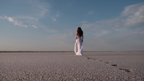 A young redheaded woman in a white dress walking on a salt lake. Woman walking along the dry bottom of a salt lake, rear view. Dry salt lake. focus on bare footprints