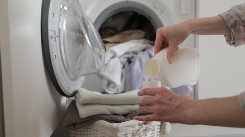Woman pouring detergent gel into cap and put into washer machine, close the door and start washer. Washer machine and clothes with wicker basket in background