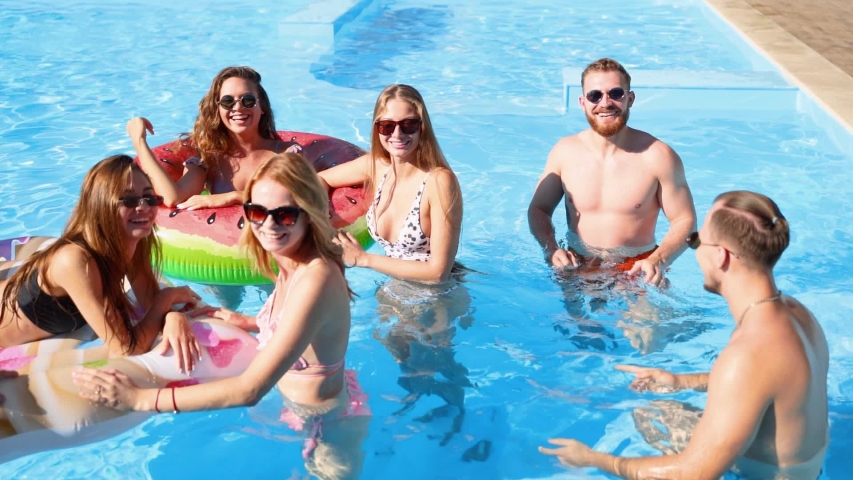 Friends have party in a private villa swimming pool. Happy young people in swimwear dancing, bonding and clubbing with floaties and inflatable mattress in luxury resort on sunny day. Tracking shot. | Shutterstock HD Video #1054318160