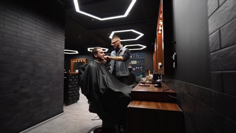Trendy barber cuts bearded man's hair with a clipper in barbershop. Men's hairstyling and hair cutting in salon. Grooming the hair with trimmer. Hairdresser doing haircut in retro hair salon. Dolly in