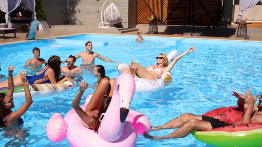 Friends chilling in private villa swimming pool, lie in the sun on inflatable flamingo, swan, floaties. Young people relax on party at luxury resort on sunny day. Bikini girls sunbathing. Slow motion. | Shutterstock HD Video #1054318205