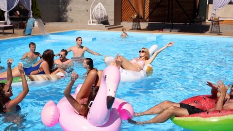 Friends chilling in private villa swimming pool, lie in the sun on inflatable flamingo, swan, floaties. Young people relax on party at luxury resort on sunny day. Bikini girls sunbathing. Slow motion.