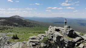 A girl stands on top of a mountain on the rocks and looks into the distance. Aerial video