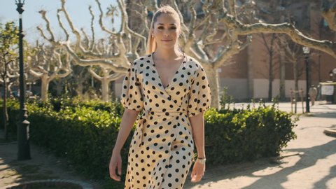 Young gorgeous woman in white polka dot dress confidently walking along beautiful city park