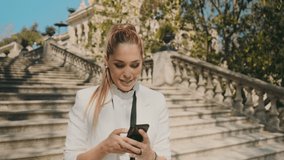 Young elegant cheerful woman happily taking selfie on smartphone on stairs in old city park over beautiful architecture. Attractive lady taking selfie outdoor