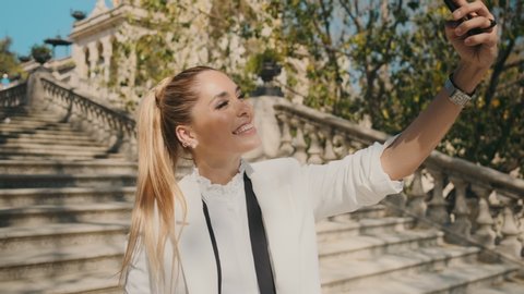Young elegant cheerful woman happily taking selfie on smartphone on stairs in old city park over beautiful architecture. Attractive lady taking selfie outdoor