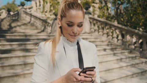 Young gorgeous elegant woman in classic suite happily using messenger on smartphone and sending selfie on stairs in old city park over historical architecture