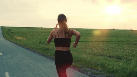 Follow-up Shot of Young Athlete Woman Running Fast down the Road, Training Hard, Getting Ready for Race Competition or Marathon. Fit Girl in Black Sportswear Jogging At Dawn along the Green Fields.