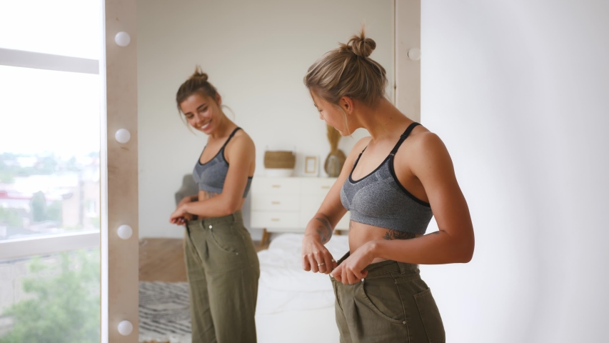 Woman is smiling, pulling jeans and showing weight loss. Standing next to mirror in bedroom. Dieting, weight loss, eating disorder concept | Shutterstock HD Video #1054326359
