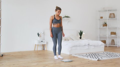 Young woman in gray sportswear standing on weight scale and rejoicing by weight loss being in white home interior. Eating disorder and weight loss