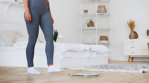 Unrecognizable girl in gray leggings standing on weight scale and dancing, happy about weight loss. Home interior. Dieting, eating disorder. Close up