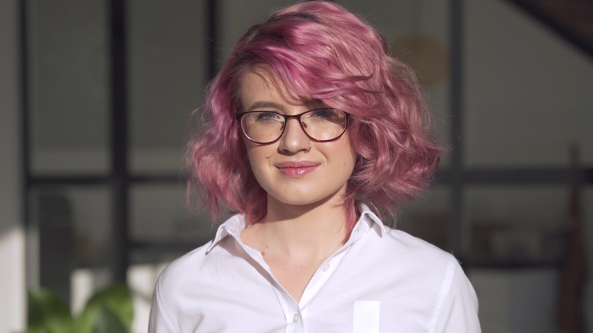 Happy young adult hipster teen girl smiling face with pink hair and piercing wearing white shirt and glasses looking at camera posing indoors in modern sunny home office. Head shot close up portrait. | Shutterstock HD Video #1054327886