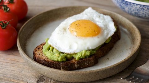 Toast with avocado and fried egg. Preparing healthy avocado toast for breakfast with mashed avocado on rye bread and sunny side up egg on top. 4K footage