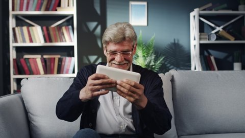 Elderly man sits on the couch in the living room and plays video games on the gadget
