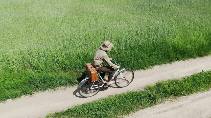 man riding on bicycle by country road in a green field, holding suitcase in a hand, funny travel concept, retro style, aerial shot from drone, top view from above, adventures, happy, creative freedom
