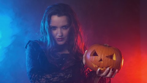 Vampiress holding jack-o-lantern. Smoky background with colored lights. Young witch in black cloak holds pumpkin in her hands. Medium shot in 4K, UHD