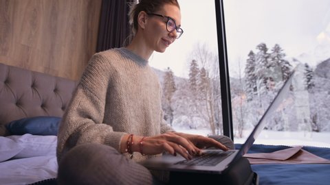 Portrait of attractive freelancer woman with glasses and a sweater with stockings sitting on a bed in an eco-house in the middle of a winter forest with a laptop