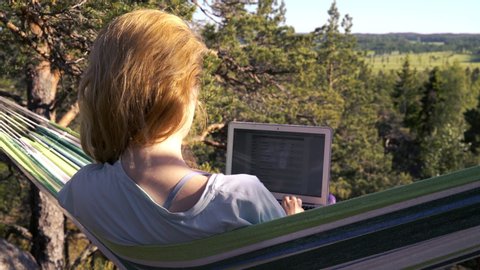 A young woman working on a laptop while lying in a hammock in the woods. Self-isolation, freelancing, remote work