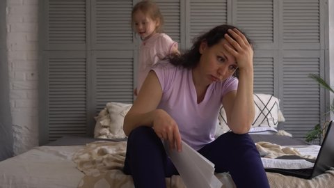 A stressed mother tries to work remotely from home during a coronavirus pandemic. A small child prevents her from working. Kid jumps on the bed. COVID-19 lockdown