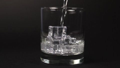 Pouring carbonate mineral water into a glass with ice cubes on a black background close-up with splashes and drops. Slow motion