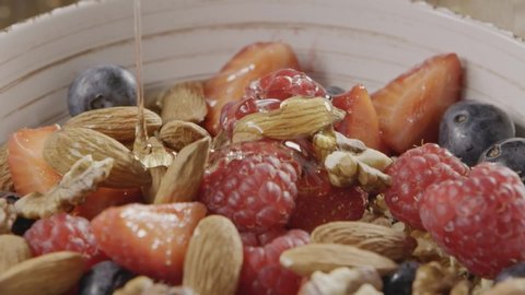 Slow video cooking healthy breakfast step by step - pouring natural organic honey into ceramic bowl with granola, nuts, mix of fresh organic fruits and berries. Slow motion, 2K video, 240fps, 1080p.