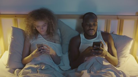 Medium shot of young family couple lying in bed with telephones in their hands. Then curly-haired young woman putting phone away, switching off light and getting ready to sleep