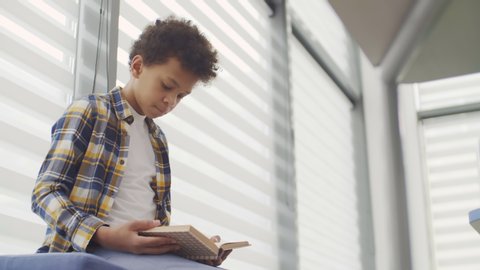 Low angle view of African boy of teenage age wearing plaid shirt and white t-shirt sitting alone on windowsill and reading book