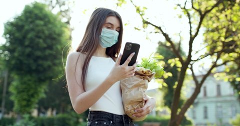 Young woman in protective mask using smartphone in the park. Worried girl searching news about Covid-19. Pandemic Covid-19 coronavirus protection. Delivery of food in quarantine