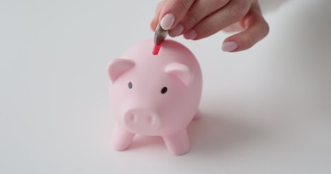 Close up young female hand putting coin in small rosy piggy bank, savings concept. Woman accounting expenses, managing monthly budget, making investment or charity, bank loan mortgage credit payments.