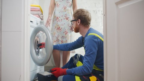 Young service man in uniform installing new washing machine and explaining woman how to use it sitting on floor in bathroom or laundry room