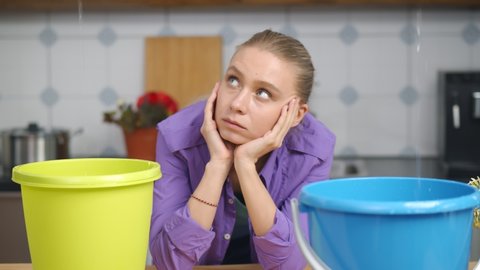 Upset woman waiting for roof repair service looking at water dropping from ceiling into plastic buckets in kitchen. Housewife waiting for plumber to fix flood