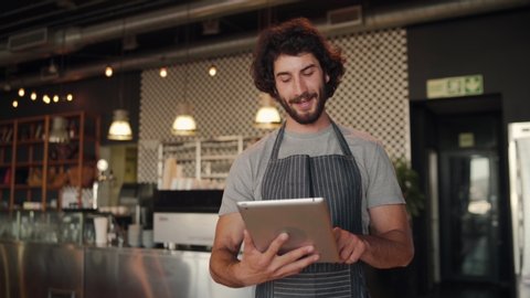 Young successful cafe owner standing in cafe wearing black striped apron using digital tablet while accepting online order