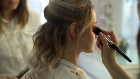 Close-up of professional hairdresser making hairstyle young attractive woman together make-up artist who applying makeup in beauty salon. Concept of beauty and fashion. Tracking shot in slow motion.