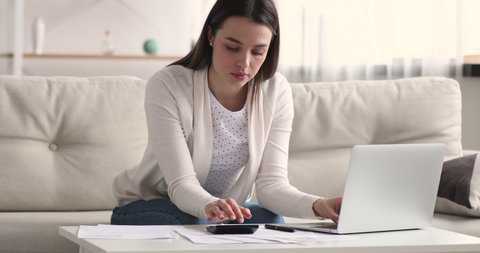 Focused serious smart young 20s woman sitting on sofa, calculating monthly budget, managing incomes and outcomes money cashflow, checking bills or rental payment, accounting taxes alone at home.