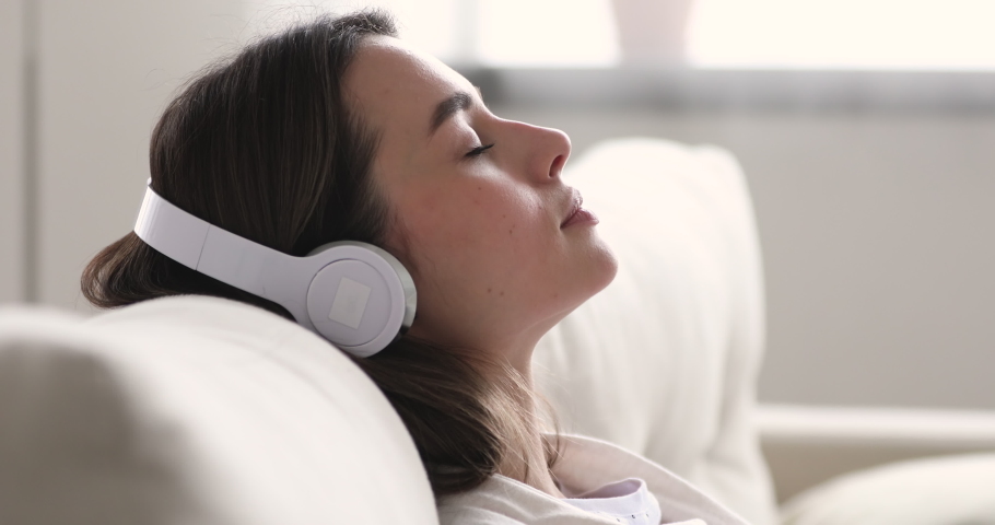 Close up head shot side view happy peaceful millennial woman relaxing on cozy couch, listening to tranquil classical music in wireless headphones, enjoying lazy weekend hobby time alone at home. | Shutterstock HD Video #1054347017