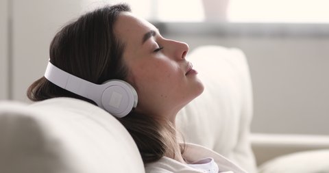 Close up head shot side view happy peaceful millennial woman relaxing on cozy couch, listening to tranquil classical music in wireless headphones, enjoying lazy weekend hobby time alone at home.