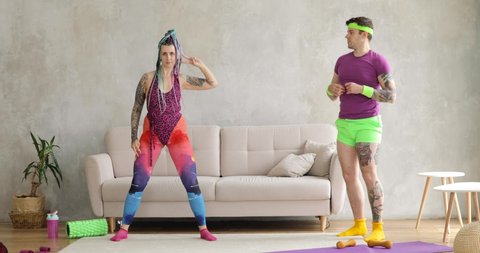 Silly dance of woman with braided hairs and funny man doing lunges sport exercise on mat at home. Joke, mem, humor, parody, joking behavior. Workout, training of couple in colourful sportswear.