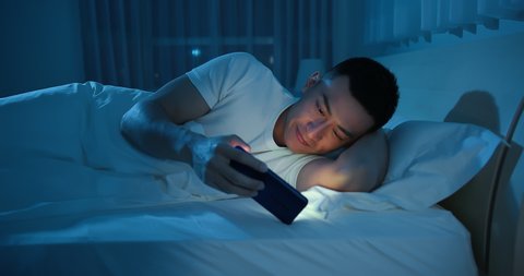 Young Asian man uses smartphone to watch video on bed at night