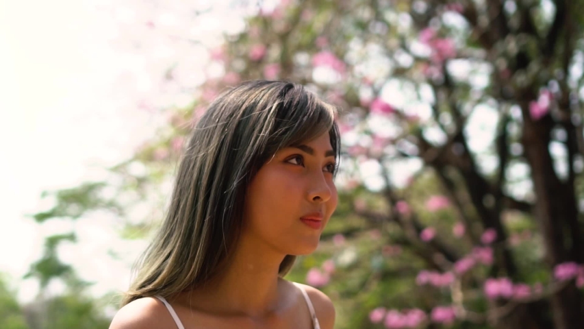 Young beautiful woman walking and looking up at flowers on tree in the park. Cute girl in summer dress smelling and feeling freedom in flower garden.