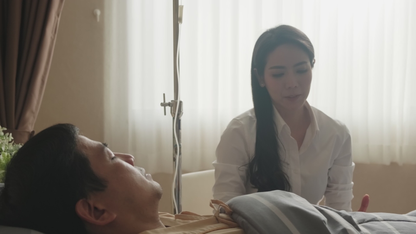 Asian patient man lay seriously ill on bed in recovery room at hospital after surgery while wife sit and look after. Woman stressed, worried about husband symptom, hold hand and bend head down crying. Royalty-Free Stock Footage #1054366016
