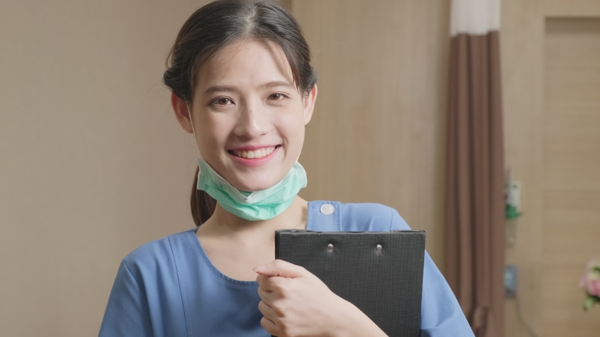 Portrait shot of Female medical nurse in medic uniform taking surgical mask off face, looking at camera and smile in slow motion shot. Beautiful Asian young woman worker is smiling in hospital.