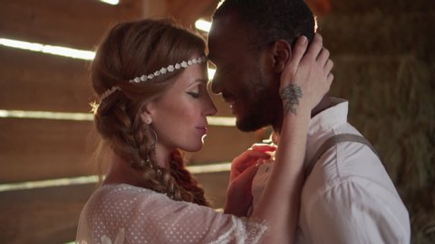 Happy Caucasian bride and African groom dressed in boho style are hugging in a barn in the sun. Close up.