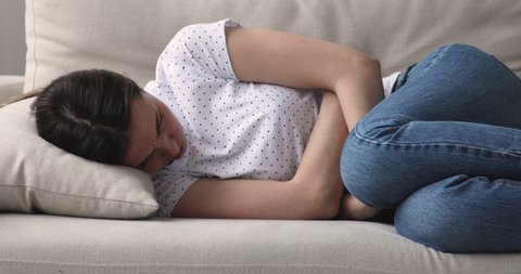 Stressed young woman lying on sofa, embracing belly, suffering from stomachache. Unhappy 20s girl having menstrual painful feelings, resting on sofa. Gastritis, abdominal or period pain concept.