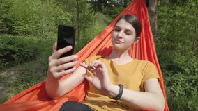 Woman in yellow t-shirt lying in a hammock and talking via video call on her mobile phone. Spending good time at the nature