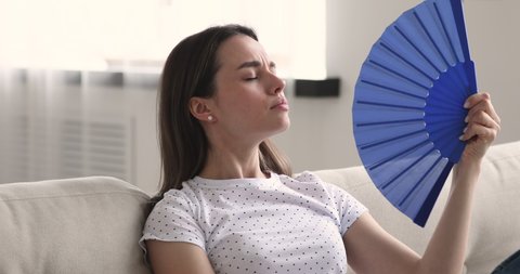 Head shot overheated young beautiful woman suffering from high temperature indoors, using paper fan. Frustrated exhausted millennial girl cooling herself, feeling hot without air conditioning at home.